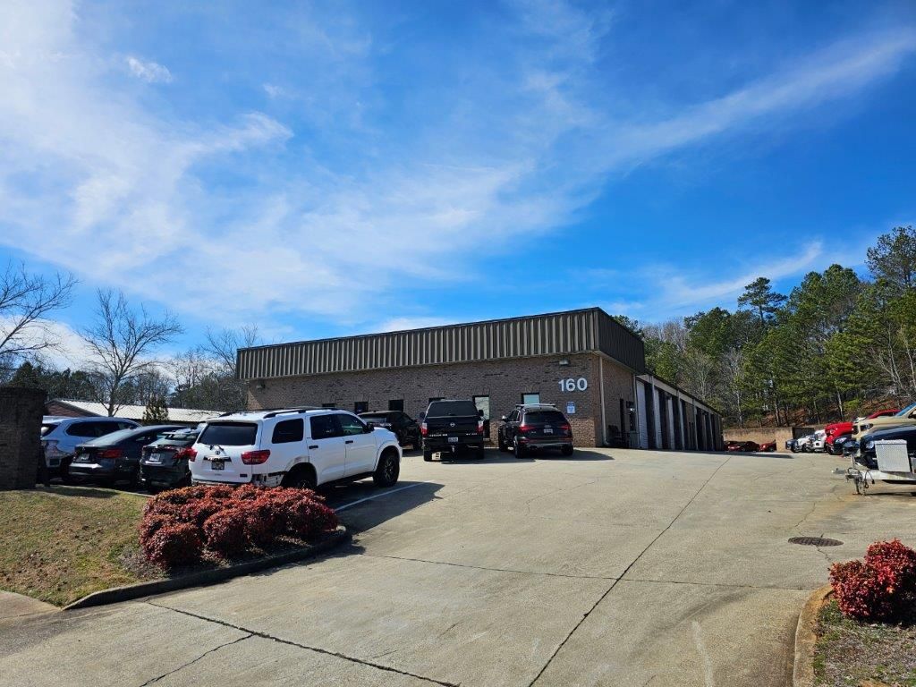 A single-story brick commercial building with the number 160, several parked cars, and a sloped driveway under a blue sky.