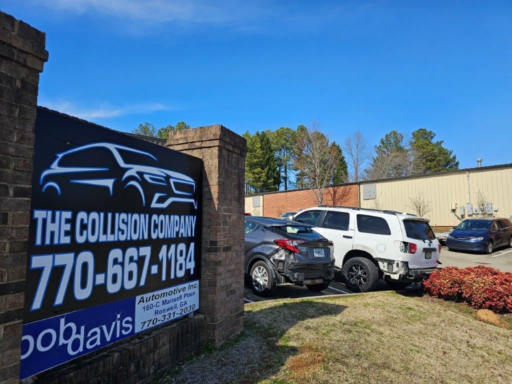 A sign for The Collision Company and Bob Davis Automotive Inc. stands by a parking lot with several parked cars in front of commercial buildings.