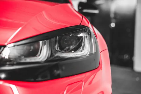Close-up of the front left headlight of a red car, showing part of the hood and bumper in a garage or indoor setting.