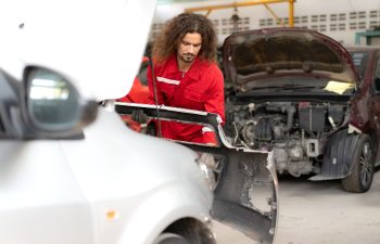 A mechanic in a red uniform removes a car bumper in an auto repair shop, with two vehicles being serviced in the background.