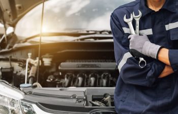 Person in coveralls holding wrenches stands in front of a car with the hood open, suggesting vehicle maintenance or repair.