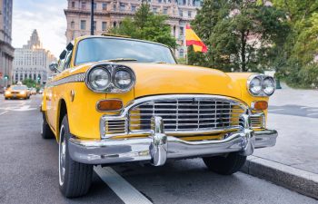 A classic yellow taxi cab is parked on a city street, with buildings and trees in the background.